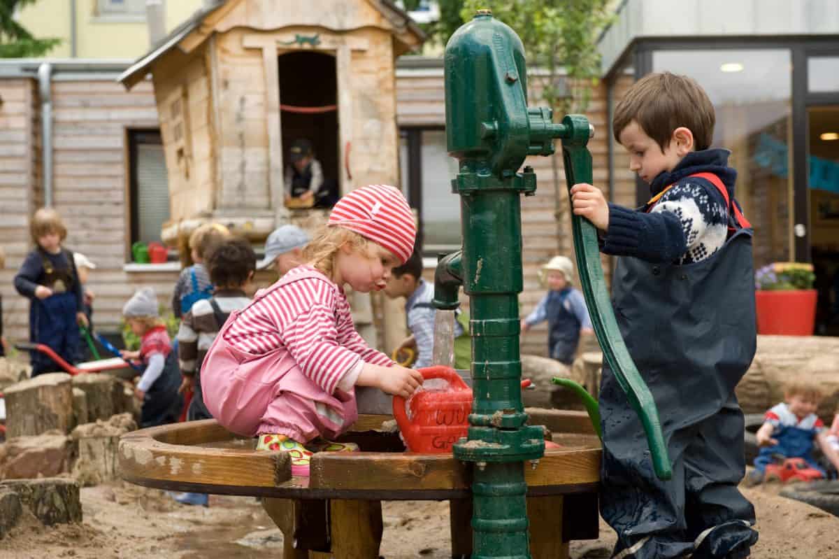 Kinder spielen mit Gießkanne an der Wasserpumpe